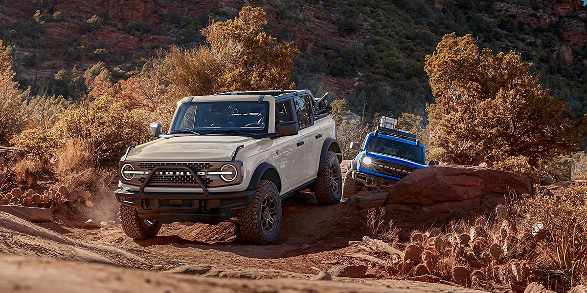 Two Ford off-road vehicles, a Bronco and Ranger, navigating rocky terrain in a desert setting, showcasing durability and adventure readiness.